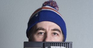 David O'Doherty. A man wearing a plaid shirt and a blue, red and white striped beanie with a bobble holds a small keyboard in front of him obscuring the bottom half of his face and all of his chest.
