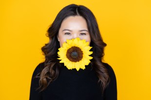 Image is a yellow backdrop behind a woman of Asian appearance with long dark wavy hair and smiling eyes, with her face partially obscured by a large sunflower.
