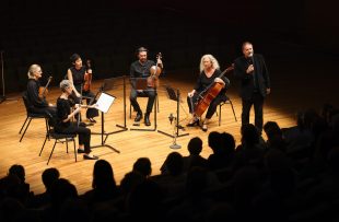 A quintet of classical musicians sit on a stage with their instruments, watched by an audience.