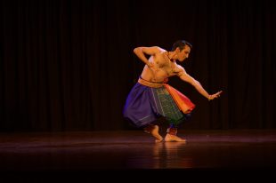 The Indian Performing Arts Convention. Image is a man of Indian appearance with dark eyeliner, shirtless and wearing green baggy pants and a red and gold sash around his waist dancing with bents knees and an outstretched hand.