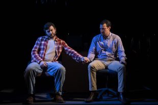 Two men sit on chairs side by side. One reaches across to hold the hand of the other.