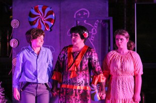 Three opera singers on a stage in front of a set depicting a candy house. A young woman plays a boy, in the middle a man in Japanese attire is in drag as a witch and on the right a woman plays a young girl. Hansel & Gretel.