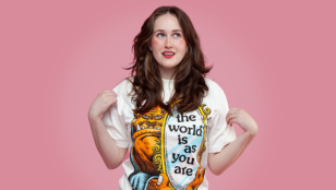 Against a pink background a young white woman with long wavy reddish/brown hair wears a T shirt with an image of a monster face and the slogan 'the world is as you are'. Grace Jarvis.