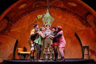 James and the Giant Peach. A man dressed in boy's shorts and shirt has two people either side turned towards him and putting their hands on him, they are dressed as insects. They are all on a stage with a big orangey red backdrop and a couple of chairs, which could be representing the inside of a peach.