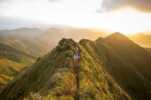 arts sector appointments. An overhead photograph of a woman jogging down the crest of a mountain ridge. More mountains stretch off into the distance, with the sun low and bright in the sky.