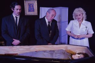 Loot. Three people on stage look down at a corpse in a coffin, a young man with a moustache, an older grey-haired man and a blonde woman in a nurse's uniform.