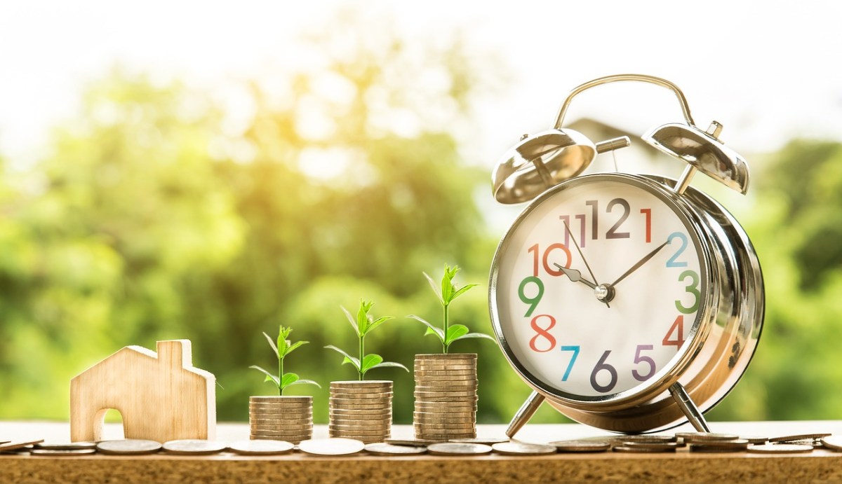 Freelance creatives. Cost of living. Image is an alarm clock, next to piles of coins and a little wooden house model, with foliage in the background.