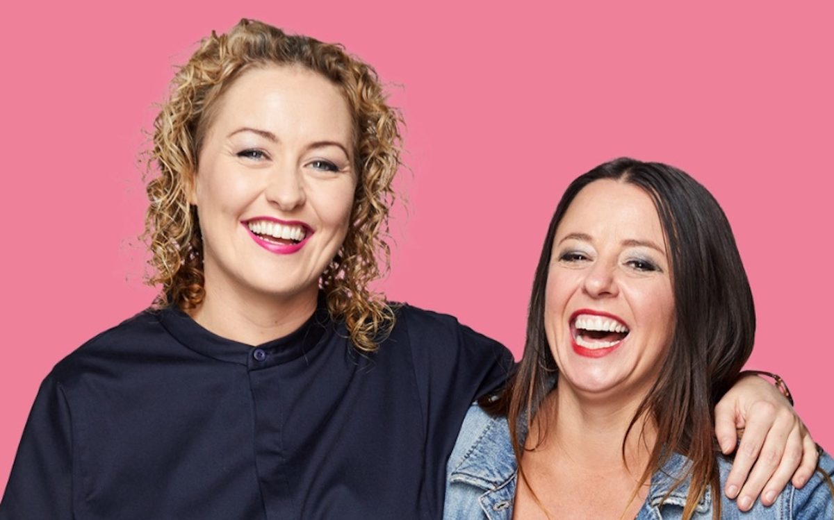 Two white women, on the left a blonde wearing all black with her arm around the shoulder of a shorter woman wearing a black singlet and denim jacket. They both have big smiles.