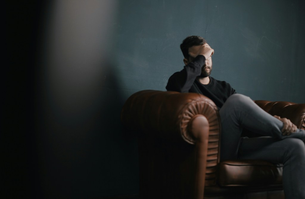 In a darkish room a man with a beard sits in a chesterfield armchair holding his forehead despondently. Self -tapes.