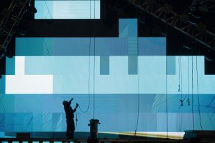 A silhouette of a person setting up a stage in a darkened space with rigging and a lit-up screen projection behind them.