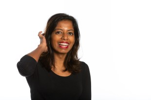A woman of south-east Asian appearance wears a black scoop neck top and smiles at the camera with one hand behind her head. Sashi Perera.