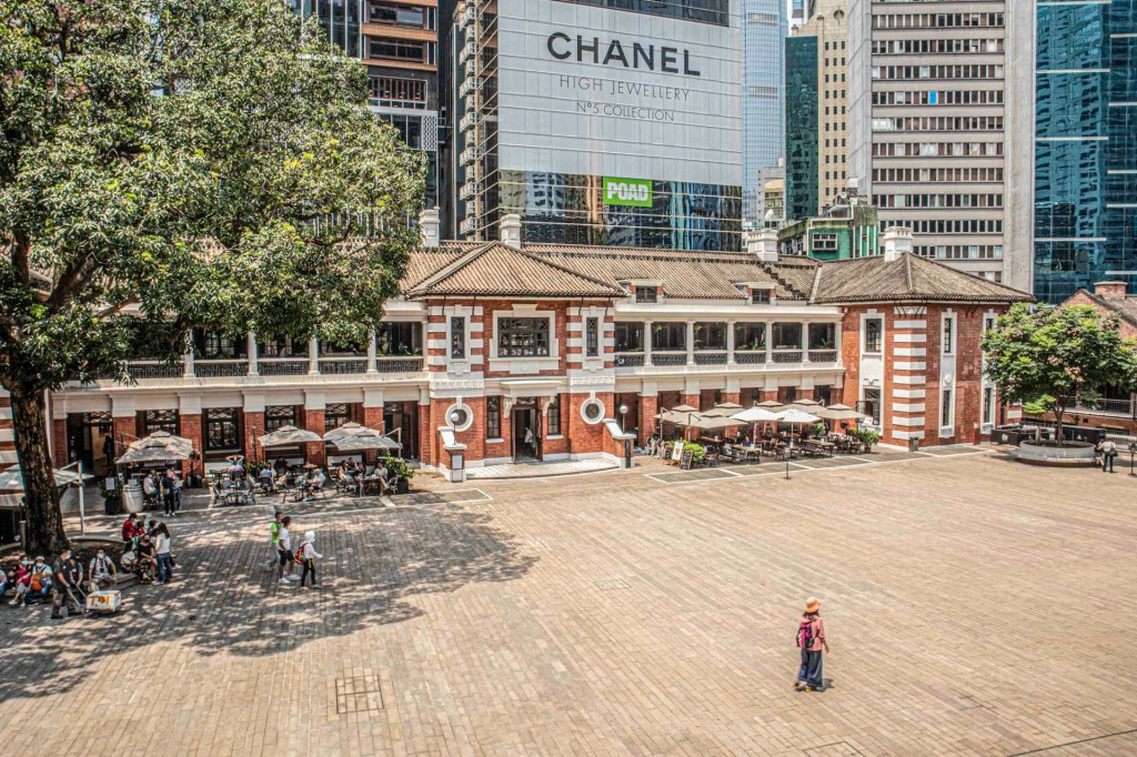 Tai Kwun, the Former Central Police Station Compound. Photo: Timmy Wong, A slightly elevated view of a spacious courtyard in broad sunlight with colonial buildings and skyscrapers nearby. People can be seem walking and sitting in the area. 