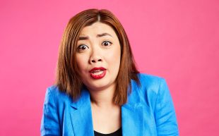 A woman of Asian appearance in a blue suit over a black scoop neck singlet has a surprised expression on her face.