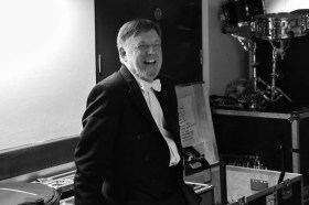 Black and white image of a smiling white man leaning against a music packing case wearing a white bow tie and formal suit.