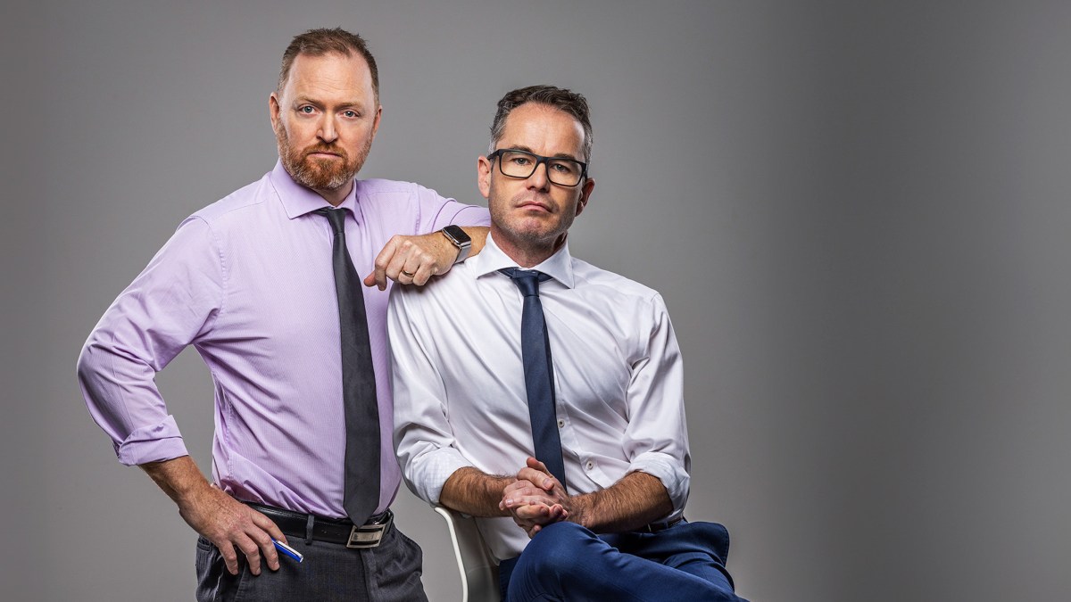 Two Caucasian men in business attire (no jackets) look seriously at the camera. One is seated, the other leaning on his shoulder.