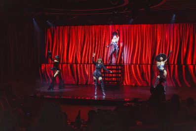 VIVA. Elegant. In front of shiny red curtains, four female dancers pose on the stage, three downstage and one on a higher platform. They are all dressed in burlesque costumes, including thigh high boots.