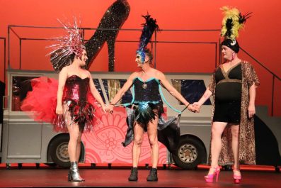 Priscilla Queen of the Desert. A couple of drag queens and a transwoman stand on stage in front of an orange backdrop and a prop bus. They are in extravagant costumes and are holding hands.