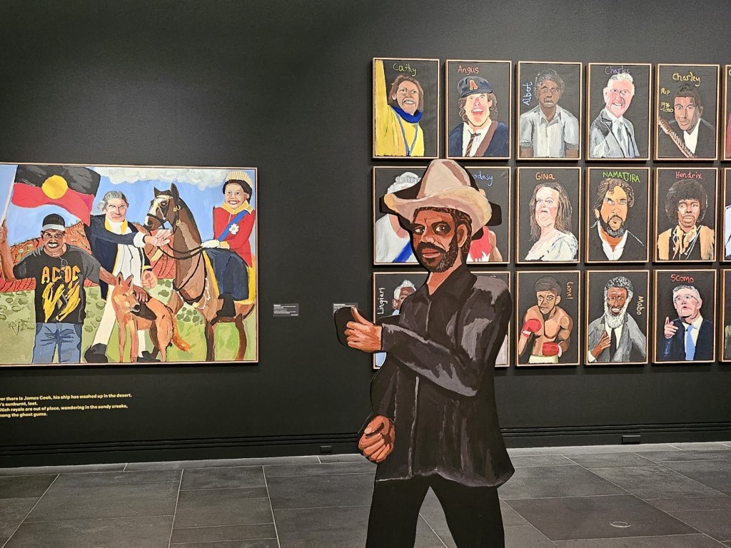 ArtsHub previously saw the work at AGSA. 'Vincent Namatjira: Australia in colour' installation view. Gina Rinehart's portrait is in the second row, third from the left. Photo: ArtsHub. A series of caricature portrait paintings on a black wall, with a cardboard cutout of Namatjira in front.