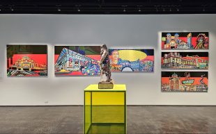 'JXSH MVIR: Forever I Live', installation view at Koorie Heritage Trust. Left to right: 'The Heart', 2016; 'Oxymoron', 2015; 'William Buckley - Maquette', 2016 (mixed media sculpture); 'Still Here', 2015; 'The Empire', 2015 (top); 'Ticket to Projection', 2015 (middle), 'Skip to my Lou', 2015 (bottom). Photo: ArtsHub. Digital illustrations of Melbourne landmarks with red, black and yellow background hang on the walls. In front of them is a small sculpture of a human figure on a bright yellow plinth.