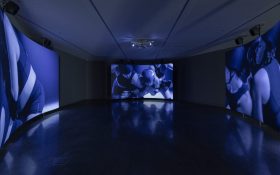 Installation view of ‘The same crowd never gathers twice’, Buxton Contemporary, the University of Melbourne, 2024. Featuring Cate Consandine, ‘RINGER’, 2024. Image: Courtesy of the artist and Sarah Scout Presents. Photo: Christian Capurro. Three channel video showing roller derby players in a blue and purple hue.