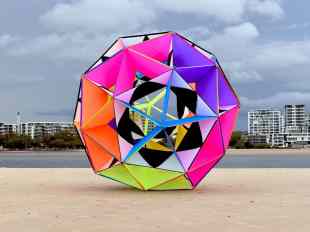 Lincoln Austin's work presents visual delights. Image is a large geometrical holey ball in bright fluorescent colours of orange, lime green, purple and pink, pictured outside with a river and some high rise buildings on the opposite bank.