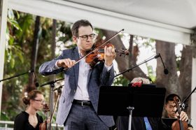 AFCM. Under an outside marquee a white man with glasses and a grey suit plays a violin from behind a music stand. There are microphones on stands. Behind him are seated a female and a male violinist resting.