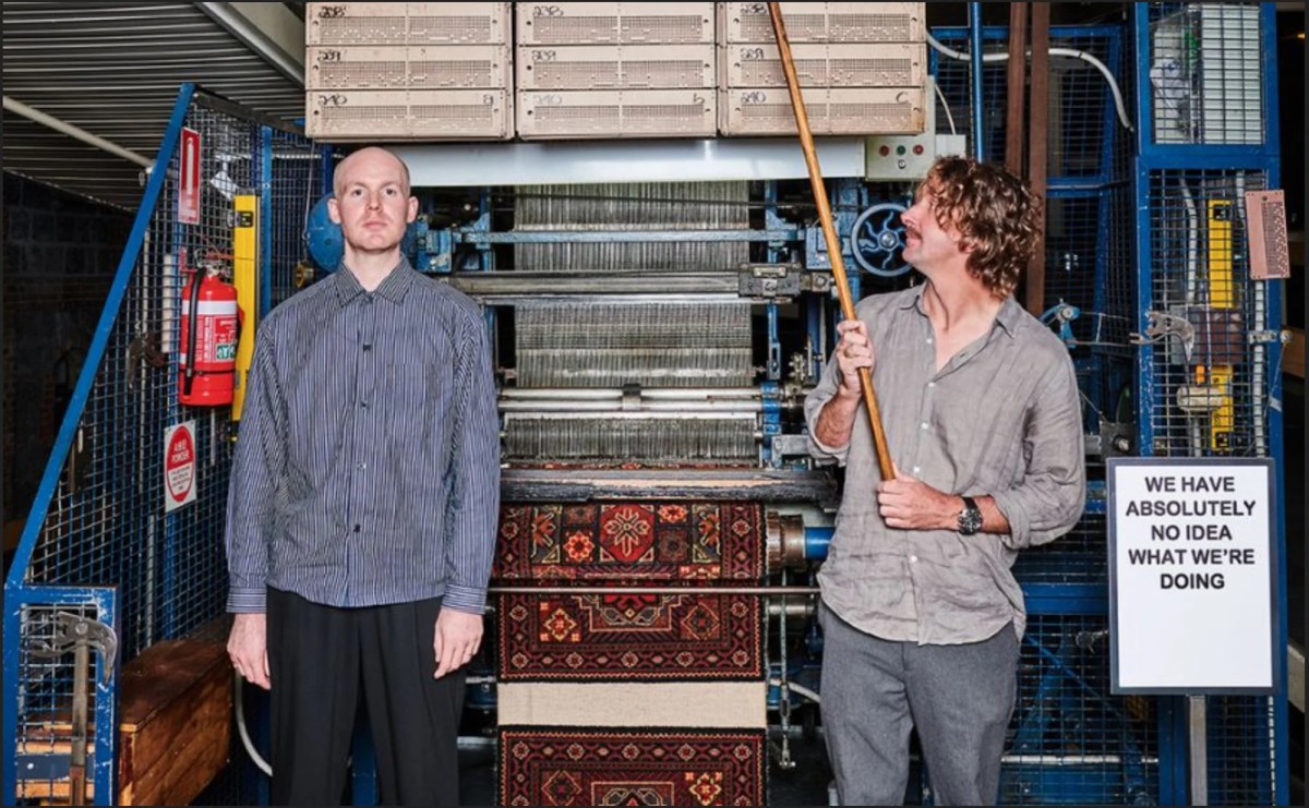 Two men wearing grey shifts standing in front of a tradition weaving rug loom. Dowel Jones.