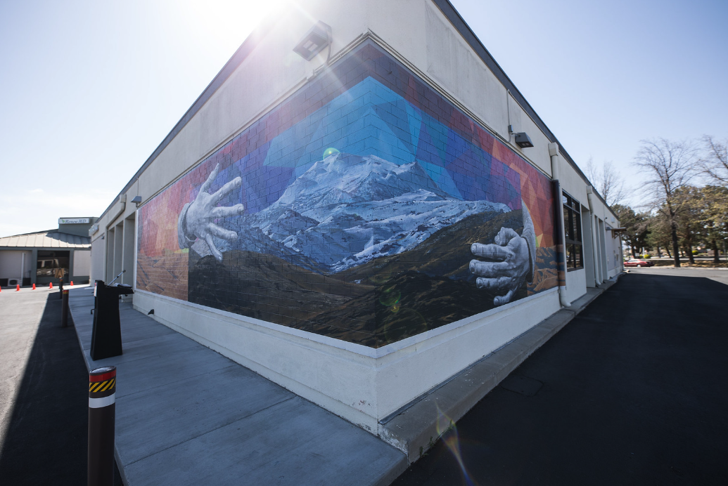 Nevada murals. From the corner of a building we can see a long wall mural that goes down either side of the building. It's an image of mountains with giants hands cradling them.