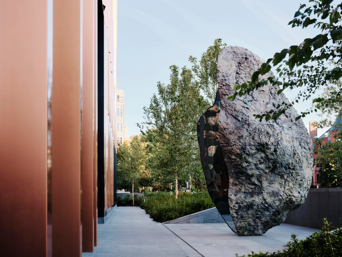 Washington DC. The outside of an architecturally designed building with brown metal standing columns along the facade on the left. On the right is a landscaped space with concrete, shrubs and trees, plus a space with a large rock shaped sculpture in the middle. 