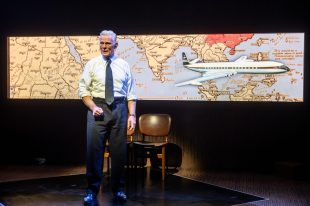 Black Box. A grey-haired man stands on a small square rostrum on a stage wearing suit trousers, a white shirt and tie. Behind him is a narrow strip screen showing a map with a large aeroplane on the right.