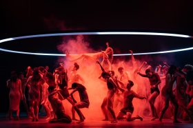 The Australian Ballet. Circle Electric. Bathed in a red glow a troupe of ballet dancers are assembled beneath a large neon hoop. The dancers are eclectic and in all sorts of shapes, one is held aloft surrounded by smoke.