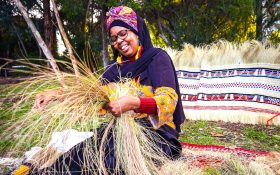 Muhubo Sulieman. Photo: Supplied. A young woman wearing a colourful purple, pink and yellow headscarf, sitting on the grass and weaving. She is in a joyful mood, smiling and working.