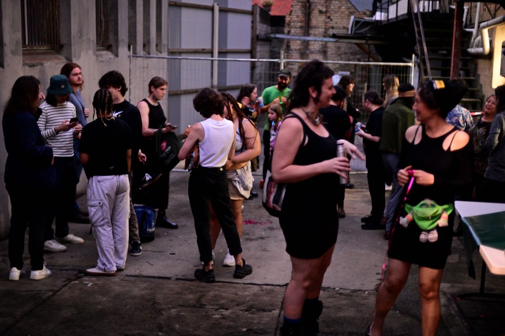 Meanjin artists. Nicola Morton (front right) conversing with a diverse crowd outside Cyber Bunker. Photo: Marek Rygielski. A crowd gathers outside a concrete alleyway in the dusk.