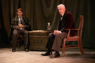 Two white men dressed in Victorian era garb sit on a stage, either side of a large theatrical costumes/props basket with a decanter on the top of it. One man is draped in a blanket and clutches a glass looking worriedly at the older grey haired man, who also looks concerned but off downstage right, clasping his hands in his lap.