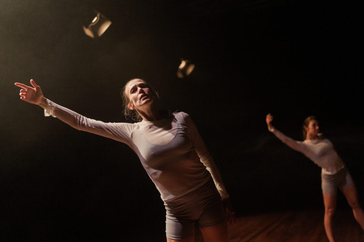 Embodied. Two female dancers on a dark stage, under two stage lights with barn doors are dressed in tight white skivvies and grey shorts, and are stretching their right arms up in the air.