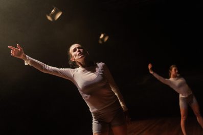 Embodied. Two female dancers on a dark stage, under two stage lights with barn doors are dressed in tight white skivvies and grey shorts, and are stretching their right arms up in the air.