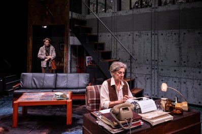 Switzerland. A theatre set of a writer's study with grey concrete walls, a two seater leather backed couch, a wooden coffee table and downstage a cluttered desk with a middle aged white woman working at a typewriter. Behind her a young white man has come through the door and looks nervous.