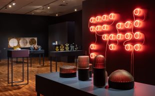 2024 FUSE Glass Prize, installation view at JamFactory Adelaide. Photo: Connor Patterson. A gallery space with black walls and timbre floor where a series of innovative glass objects are placed on several narrow tables. A large red neon work is installed on a wall to the right.