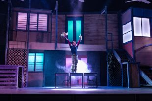 Fourteen. A stage set of a two-storey building, in the middle of the stage standing on the middle of three desks is a young man holding a bunch of flowers and throwing both arms in the air extravagantly.