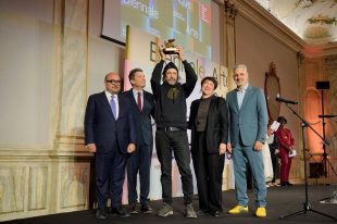 Golden Lion. Four men and one woman stand on a stage with a Biennale Arte backdrop. The man in the middle is First Nations and wears a beanie; he is holding the Golden Lion aloft.