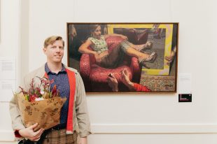 Hornsby Art Prize. Image is of a blond haired white man in his 30s holding a bunch of flowers and standing in front of his painting, which depicts a young woman lying across a red armchair with big gold hooped earrings, rolled up brown trousers, a green and white crop top and high heeled brown and white zebra print shoes, with various hands and arms reaching in from the edges of the frame holding balloons, striking a match, making gestures at her. She looks off to the right.