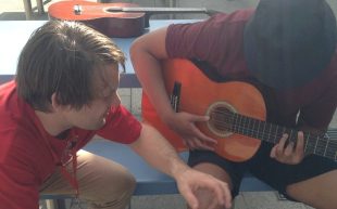The ACMF music program at Cobham Youth Justice Centre NSW. Photo: Supplied. Two people in red polo shirts sitting outside on a bench. One is holding a guitar with the head down while the other appears to be teaching.