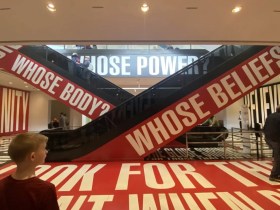 Washington DC. A gallery space with large red and white installation of phrases, including WHOSE BODY? and WHOSE BELIEFS? diagonally up the sides of escalators and WHOSE POWER? on the back wall. Another incomplete sentence is on the floor, and a young boy looks on with his back to us from the bottom left corner of the frame.