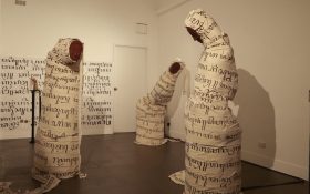 Installation view of ‘Anugerah: (Bestowed)’ at HAIR ARI. Photo: Courtesy of HAIR ARI. Three figures with no limbs wrapped with cotton straps with Javanese text, each of them is wearing a clay mask. They are inside a gallery space with dim warm lighting and more Javanese text on the wall.