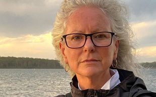 Rosemary Laing (1959-2024). Photo: Courtesy of Tolarno Galleries. A Caucasian woman with flowing grey hair, wearing a black-rimmed glasses and a black windbreaker. The background appears to be a lake.