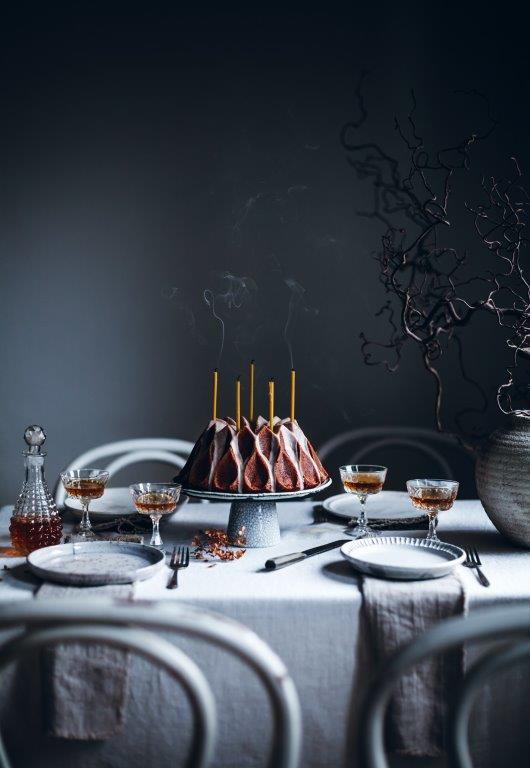 A photograph of a formally set table, with an ornate cake in the centre, half drunk glasses of wine, a decanter, plates and runners. The Art of the Table.