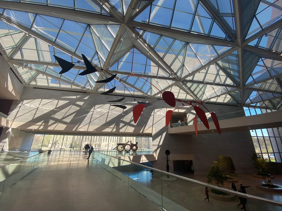 Washington DC. An expansive architectural atrium space with a glass roof filled with crisscrossing beams and girders. Outside can be seen blue sky and buildings. In the mezzanine is a large abstract sculpture from 1976.