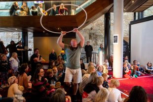 ROOKE perform Nimble at Theatre Royal Hobart. A balding man with a beard holds a hoop up over the heads of an audience of children and parents sitting on the floor. He wears a green T shirt and white shorts.