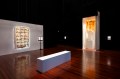 Image is a dark gallery with a shiny wooden floor and a white box seat. On the walls are two 3D pieces, one a cupboard like work and the other comprising abstract faces.