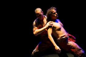 Two Aboriginal men dance together, both dressed in orange prison overalls. One, fully dressed and with a shaved head, supports the other man, who has curly hair and is shirtless, his overalls tied around his waist.